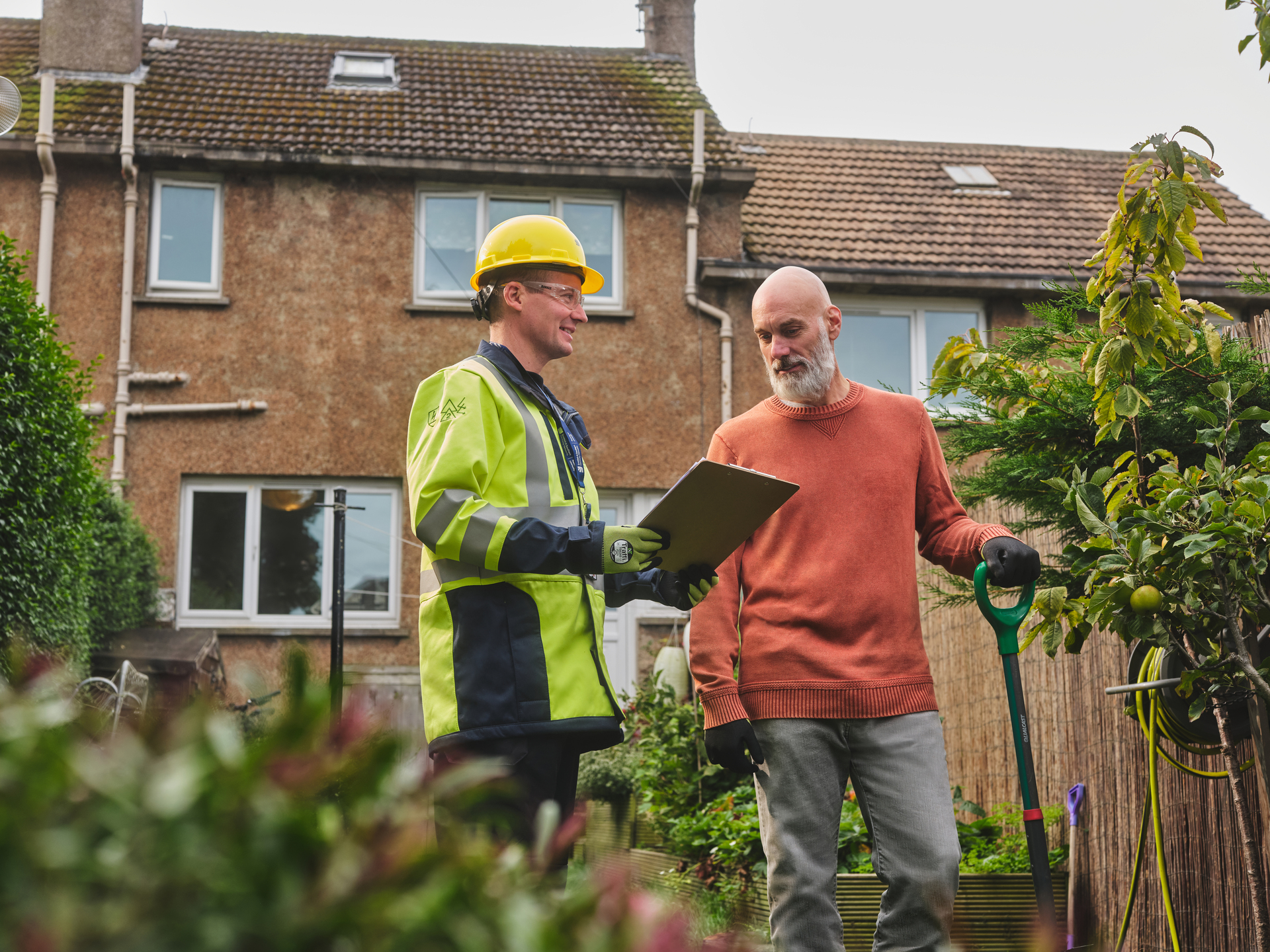 SSE engineer in full bright yellow PPE standing talking to a customer in a garden with a house in the background