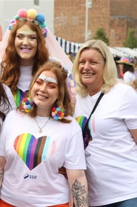 Three SSE female colleagues in SSE-branded Pride tops smiling with colourful accessories on