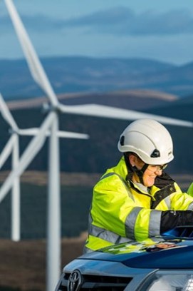 A female engineer leaning on a blue van wearing a hard hat and high visibility jacket with an onshore wind farm in the background