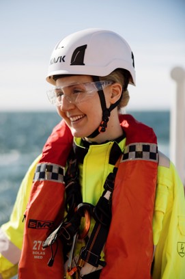 The top half of a female engineer smiling wearing a hard hat, safety glasses, high visibility jacket and life jacket with the sea in the background
