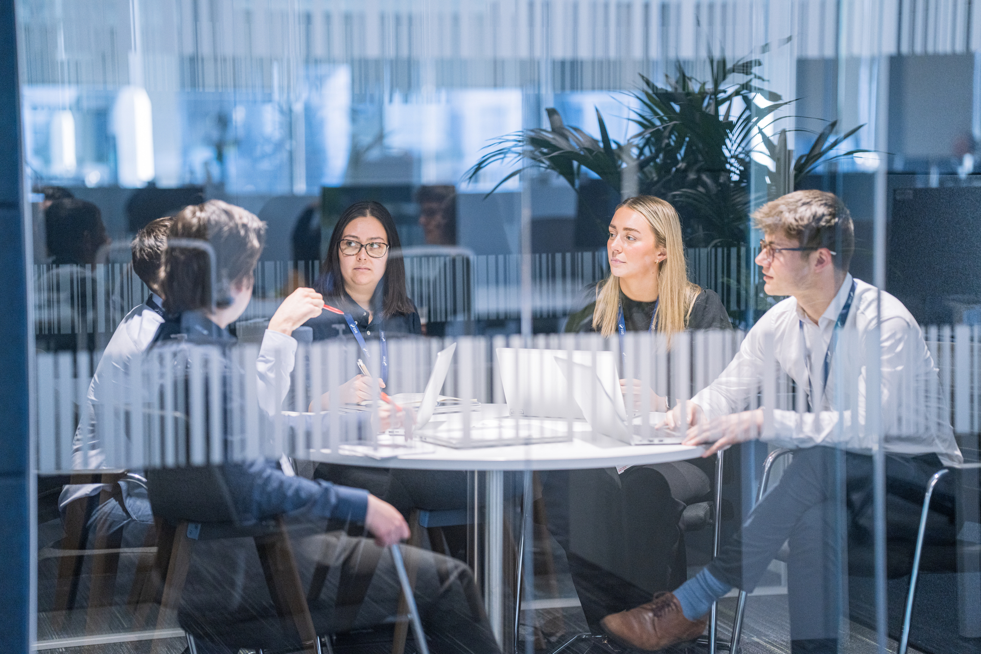 A group of 5 people sat around a desk in a small meeting room having a discussion