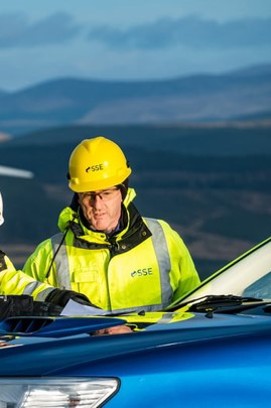 Male engineer in a hard hat and SSE branded high visibility jacket standing behind a van with rolling hills in the background