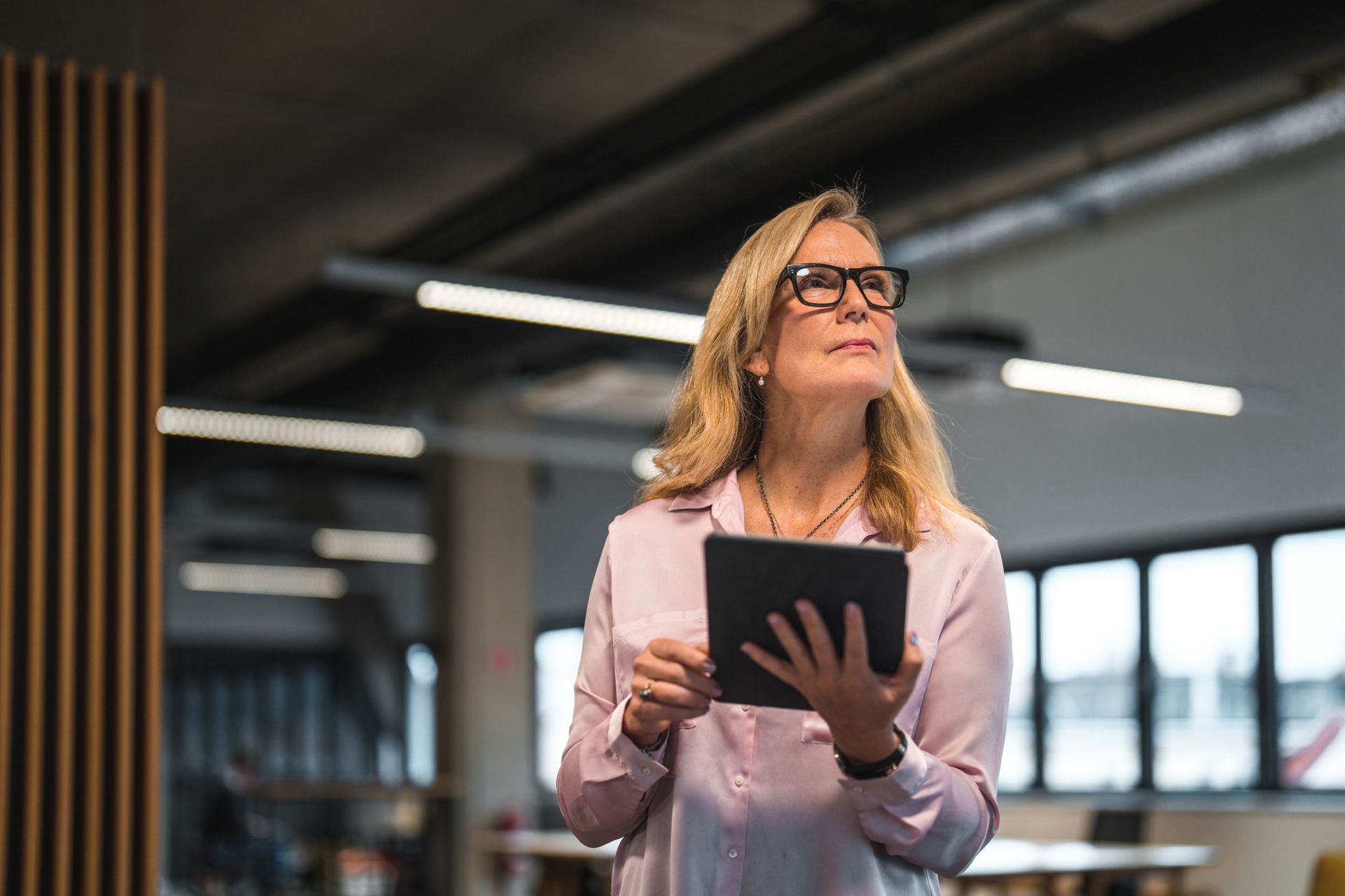 A female SSE employee with blonde hair, a pale pink shirt and oversized glasses is standing in an office holding a tablet whilst looking at something whilst showing an expression on concentration