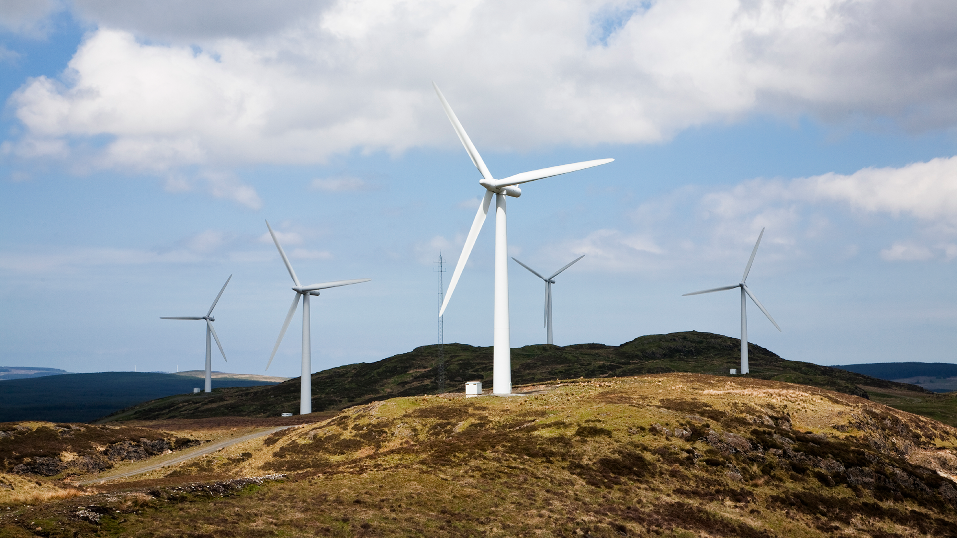 6 onshore wind turbines on the top of a green and brown hilly area and a blue sky with white clouds in the background